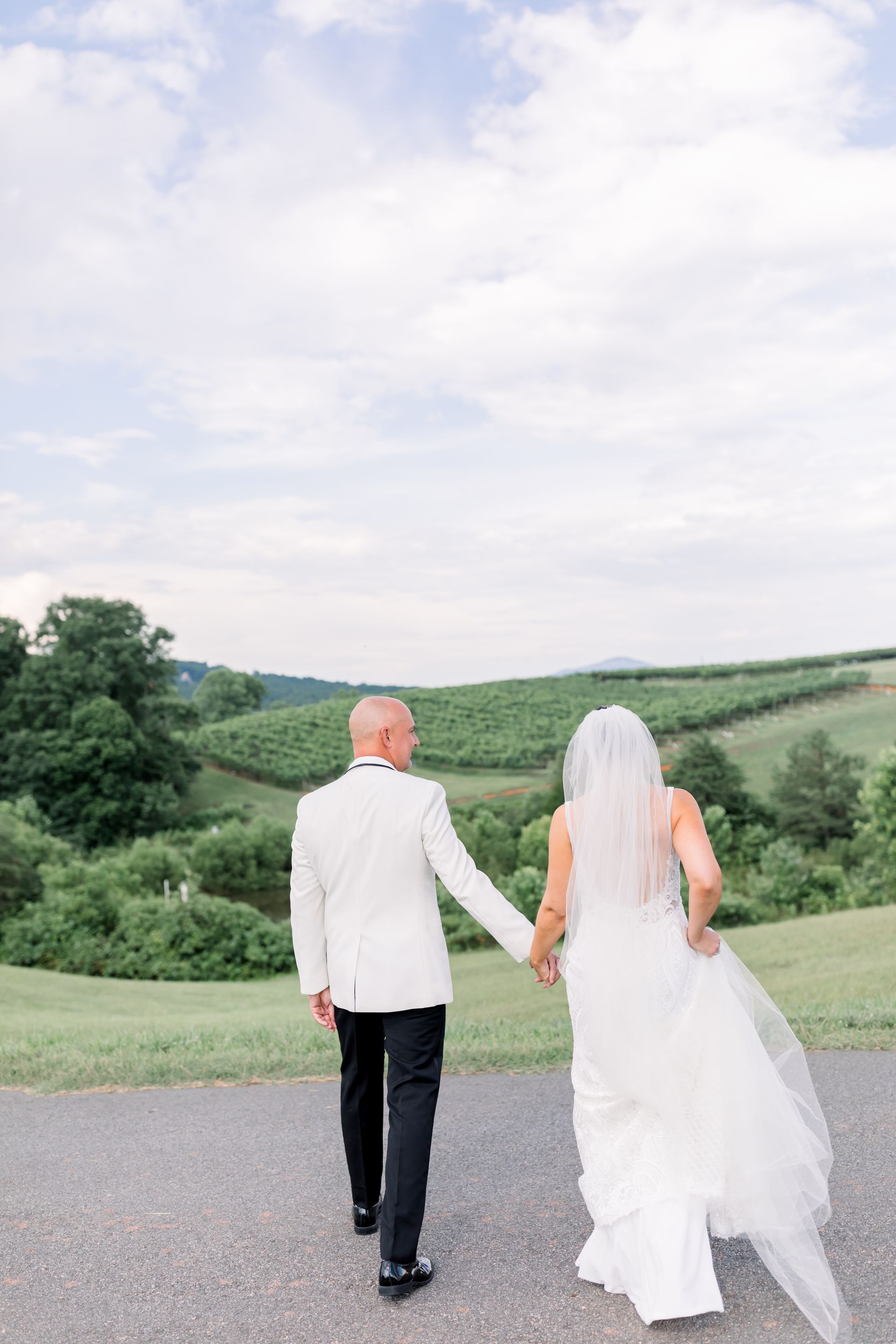 Couple in wedding attire walk away, holding hands, down a path; background of trees and fields.