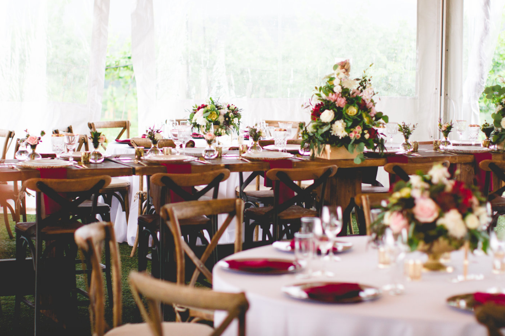 Wedding reception tables set with flowers, burgundy accents, and wooden chairs inside a tent.