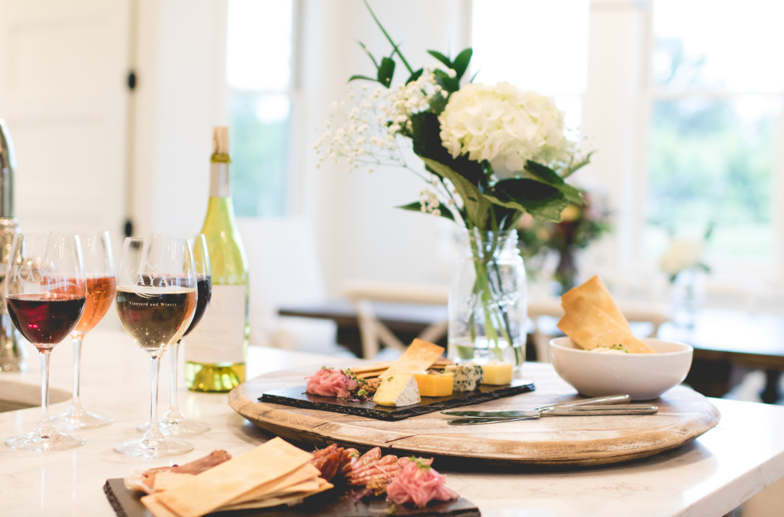 Wine and cheese board with white flowers on a counter in a bright room.