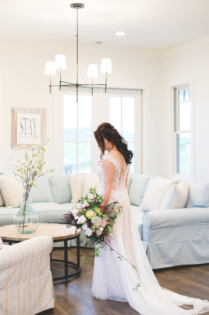 Bride in wedding dress, holding bouquet, inside a bright living room.