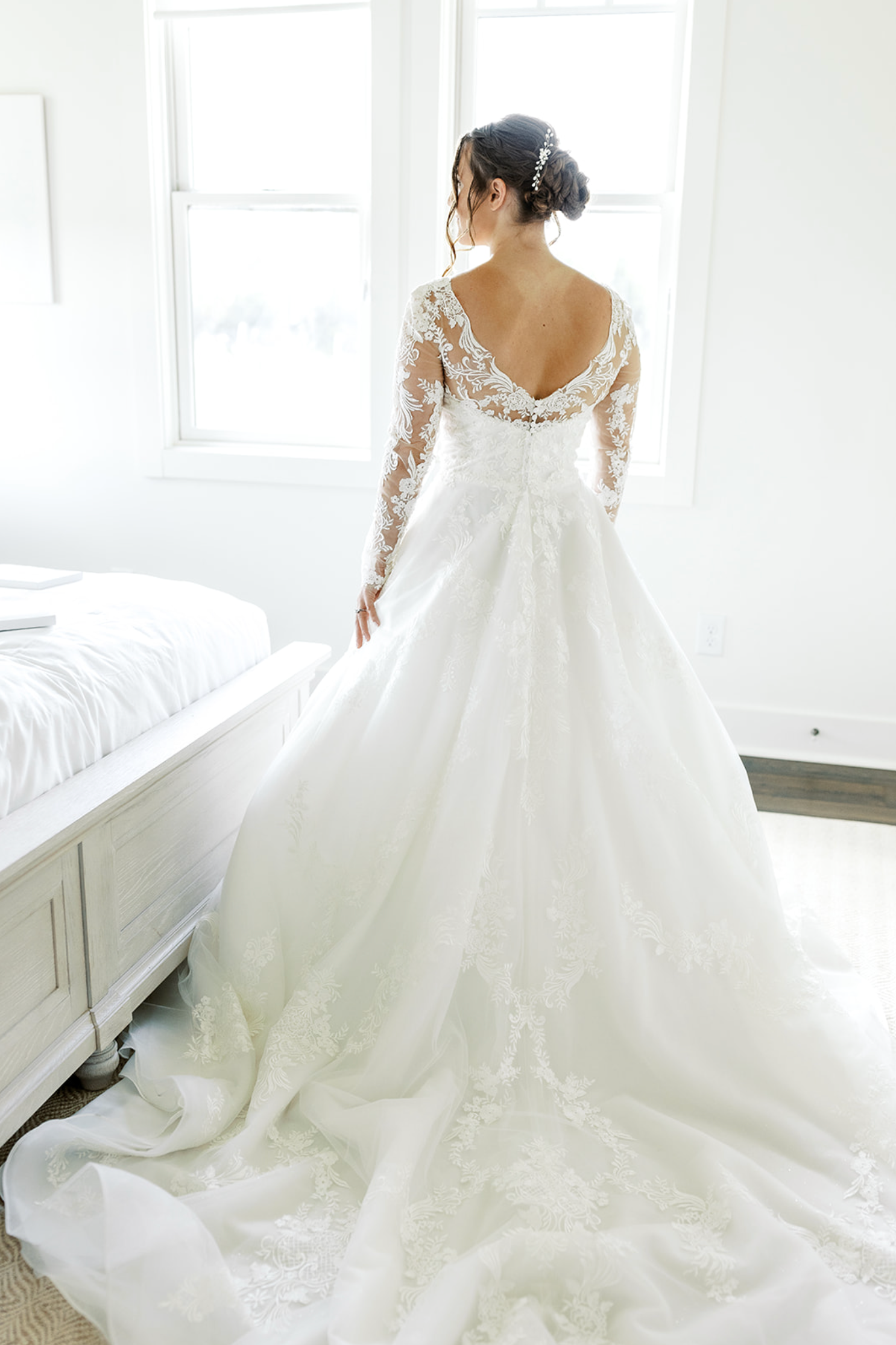 Bride in wedding dress, standing near window, back to camera, dress train on floor.