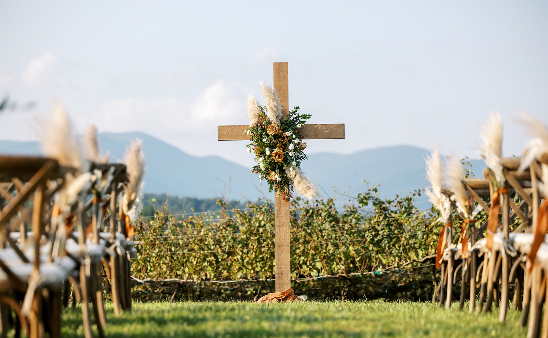 Wooden cross decorated with flowers at an outdoor wedding ceremony, rows of chairs lead to the cross, mountains in the background.