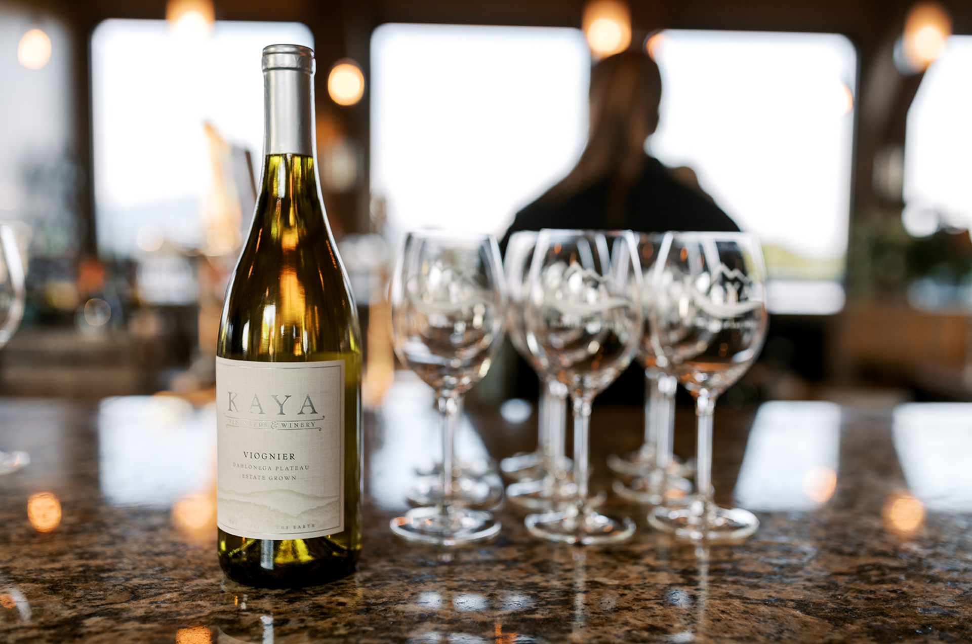 Bottle of white wine and wine glasses on a countertop with a blurred figure in the background.