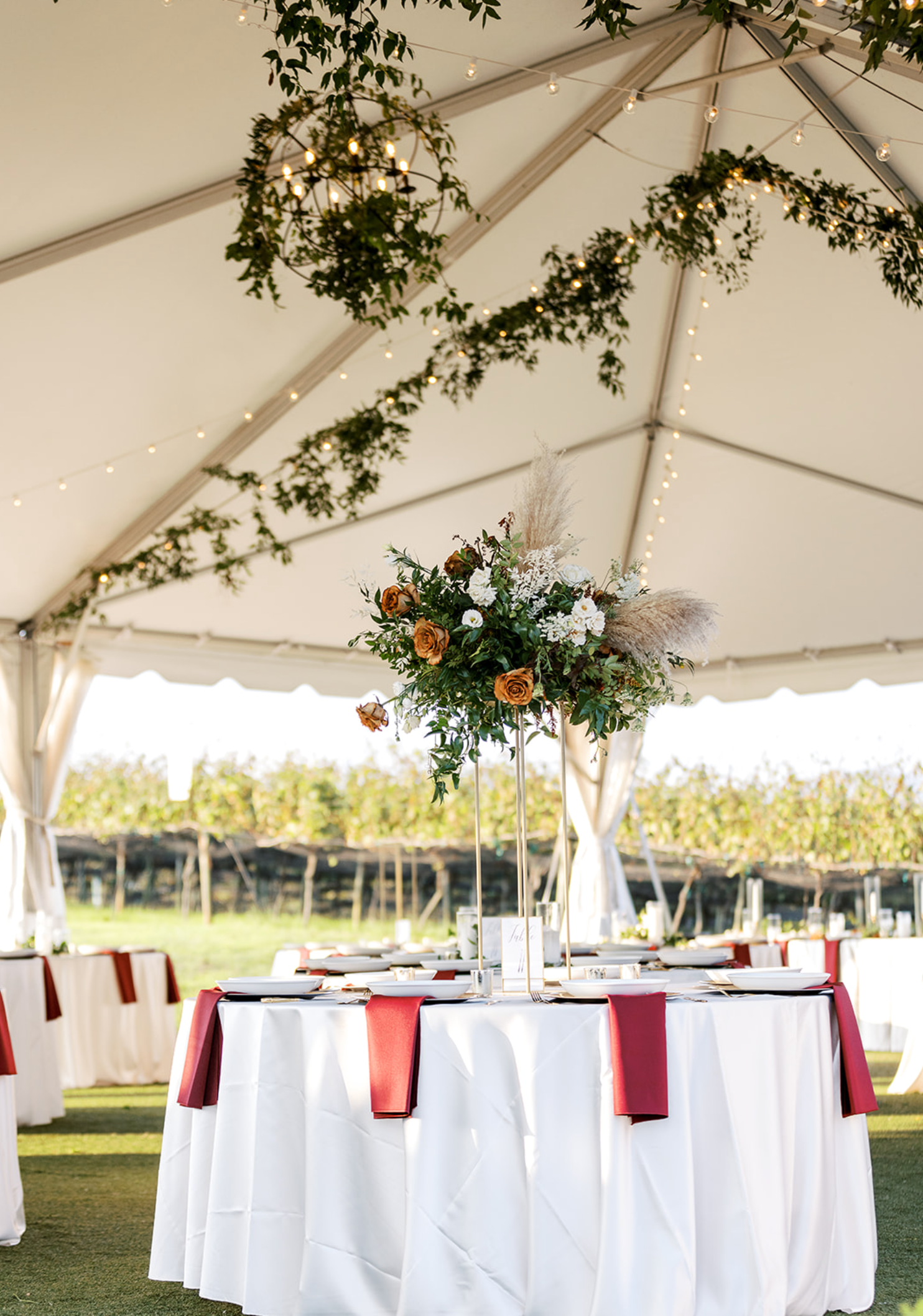Wedding reception table with floral centerpiece under a tent adorned with greenery.