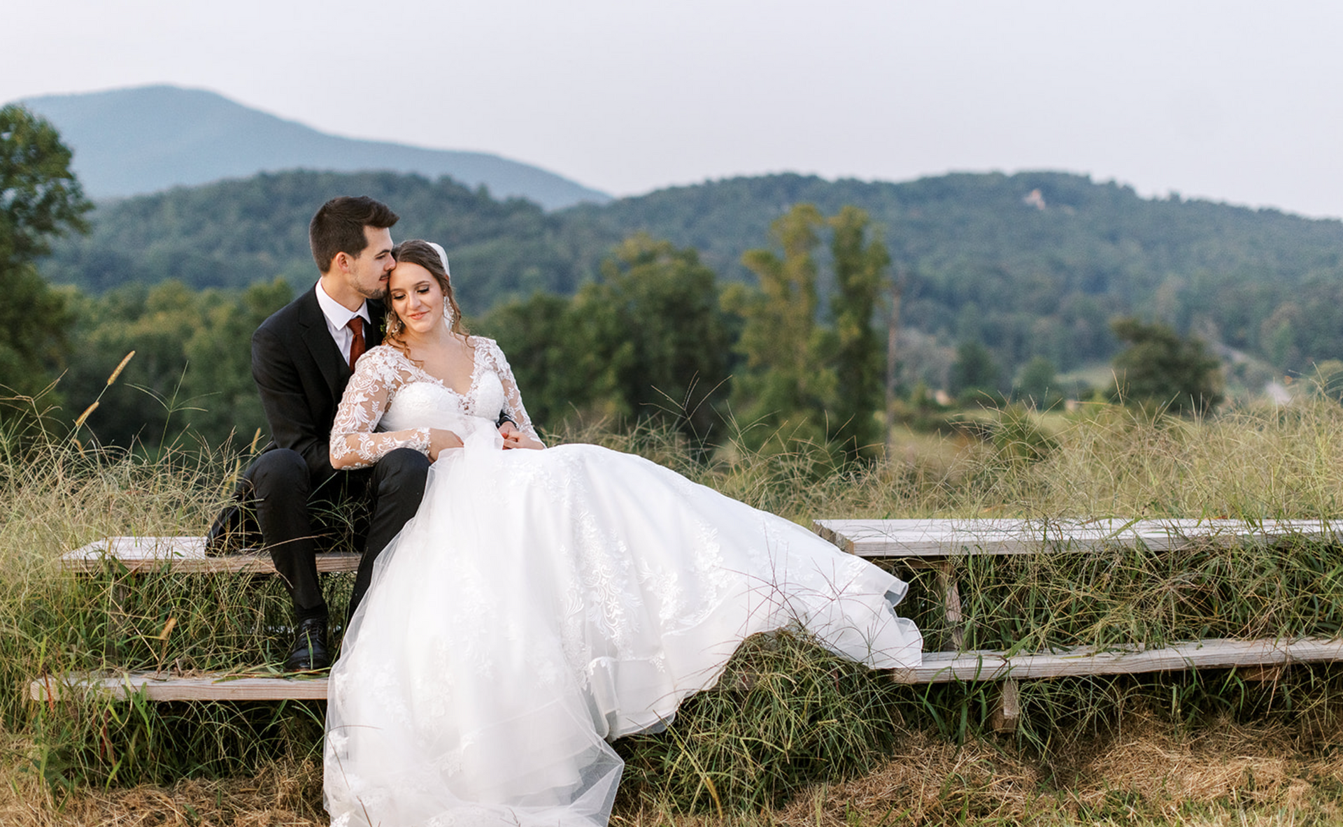 Bride and groom on a wooden bench, mountains in the background. Bride in white dress, groom in black suit.