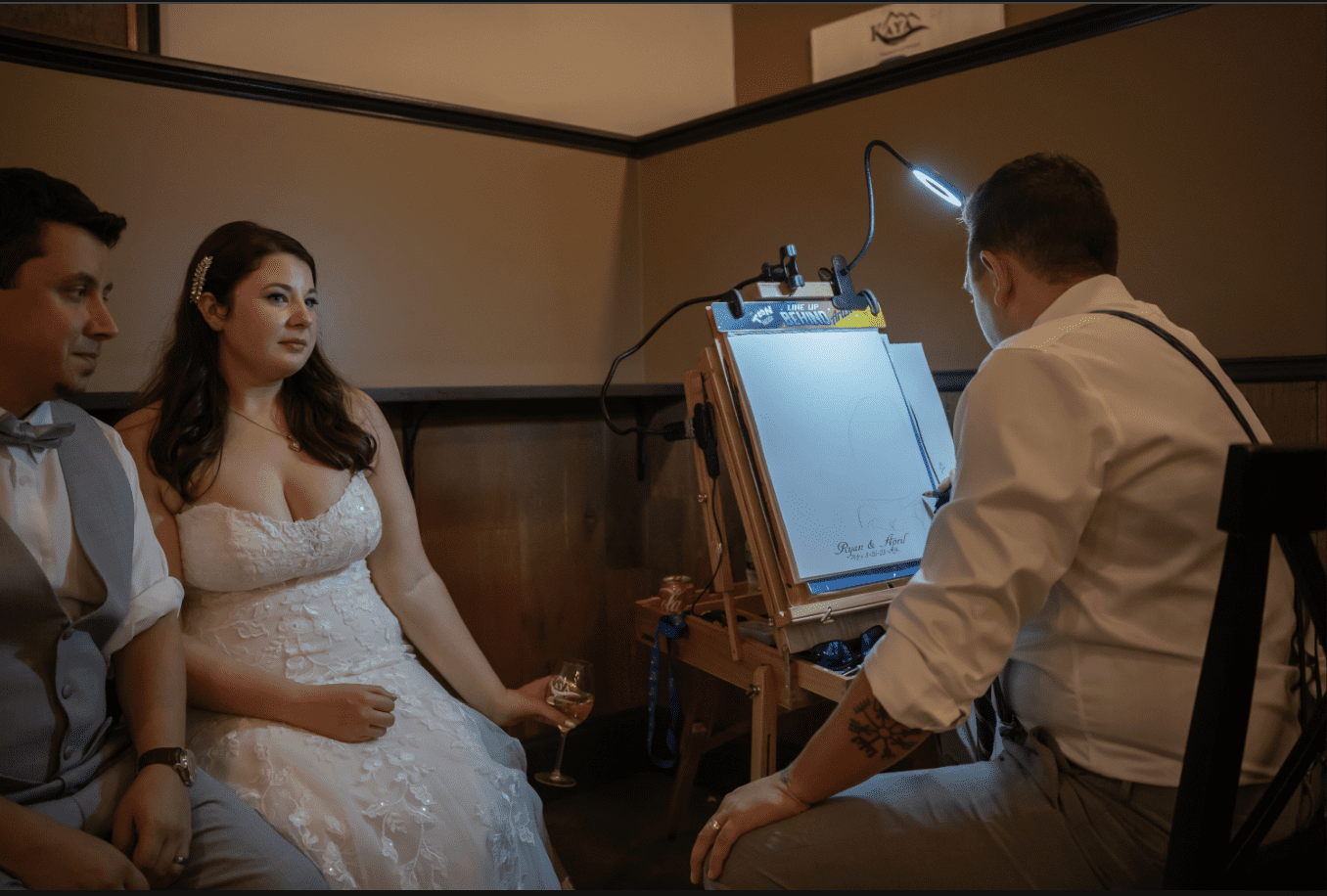 Bride and groom pose for a caricature at their wedding reception. The artist works with a lighted easel.
