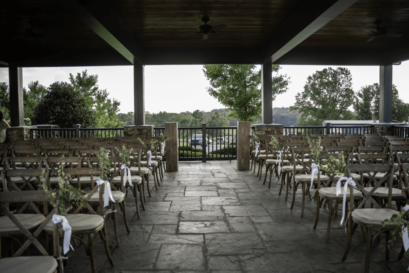 Wedding ceremony setup under a covered patio; rows of wooden chairs with white bows, overlooking a lake.