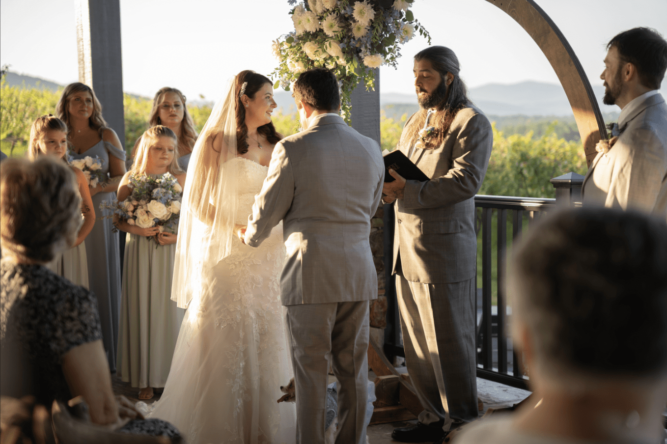Wedding ceremony with couple exchanging vows outdoors; bridesmaids, officiant, and guests present.