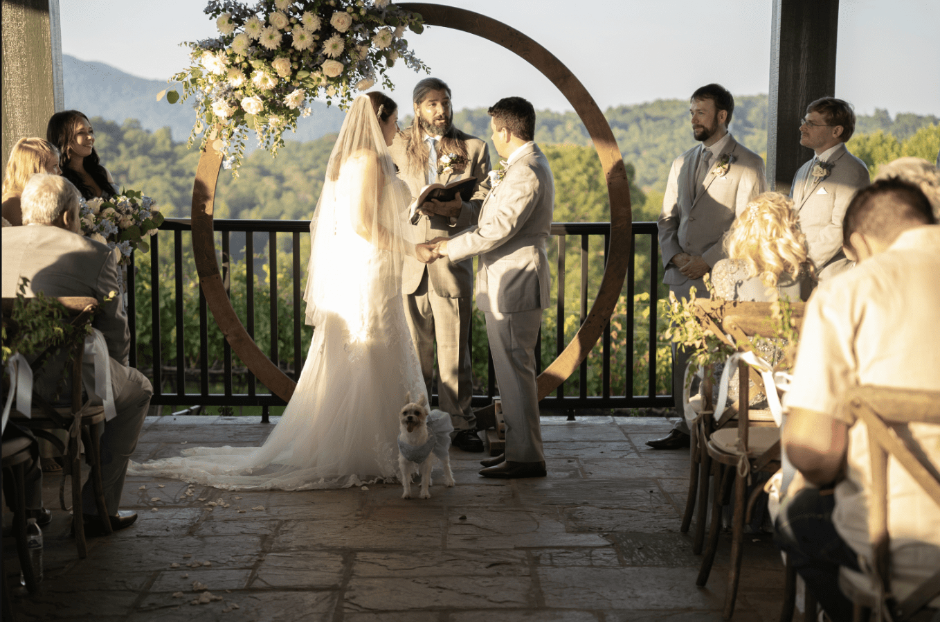 Wedding ceremony: Couple exchanging vows under floral arch, with attendants and mountain view.
