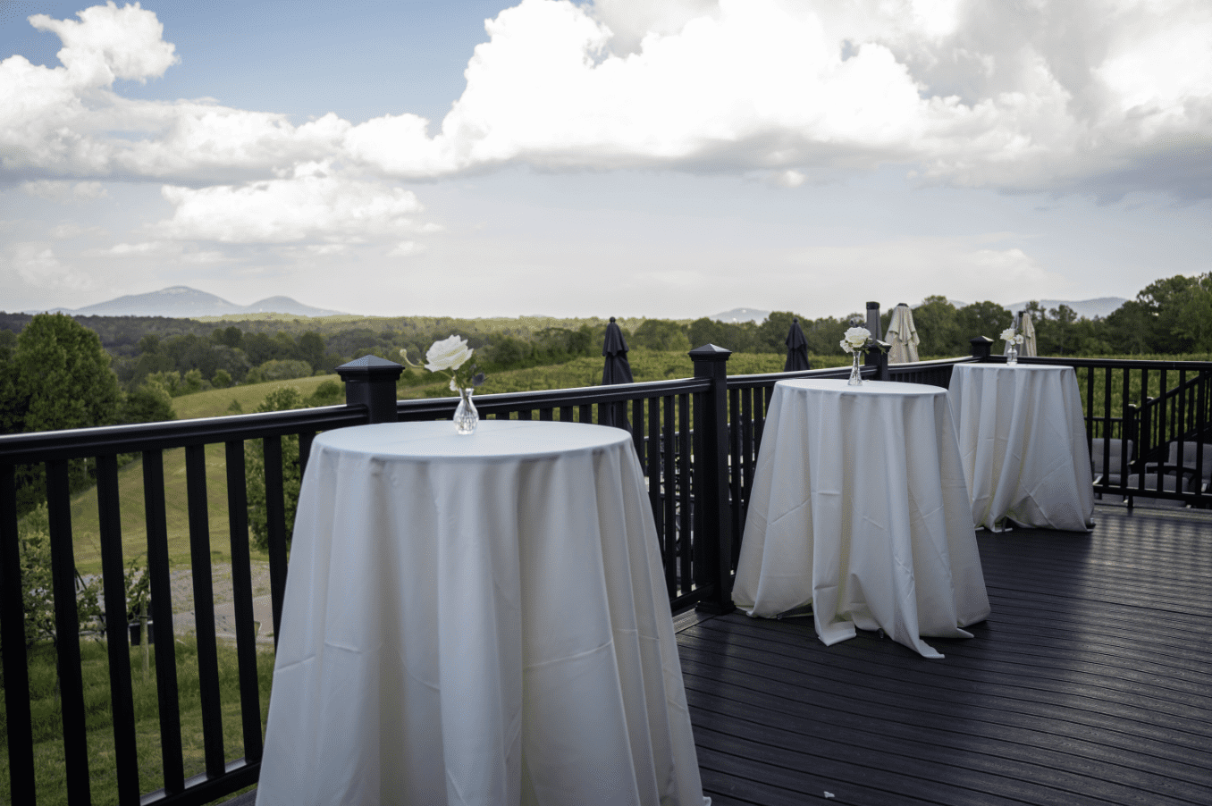 Cocktail tables on a wooden deck overlooking a scenic landscape. Cloudy sky.