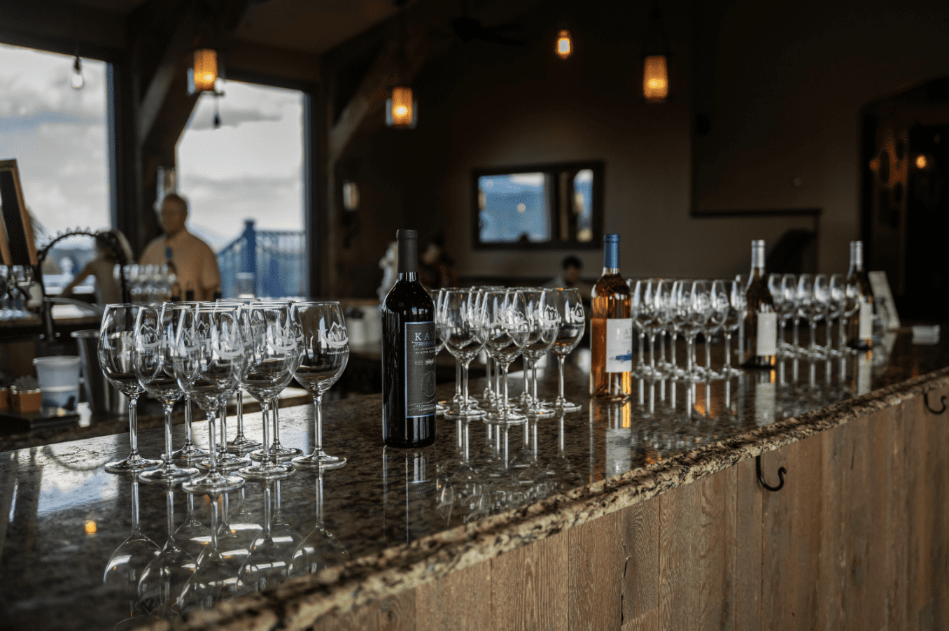 Bar with rows of wine glasses and bottles, likely for a wine tasting. A man stands in the background.