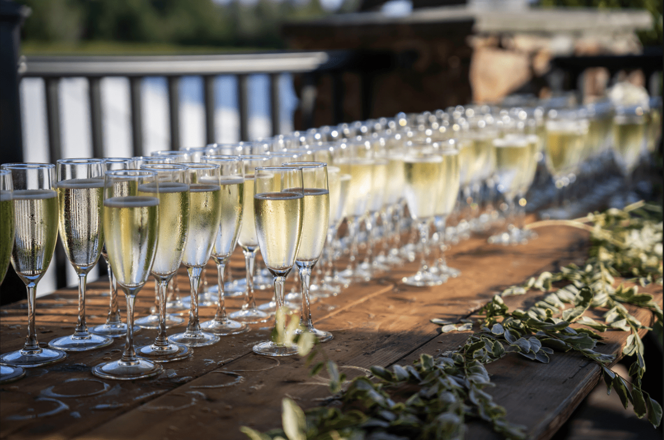 Champagne flutes lined up on a wooden table, ready for a toast. Greenery decorates the edge.