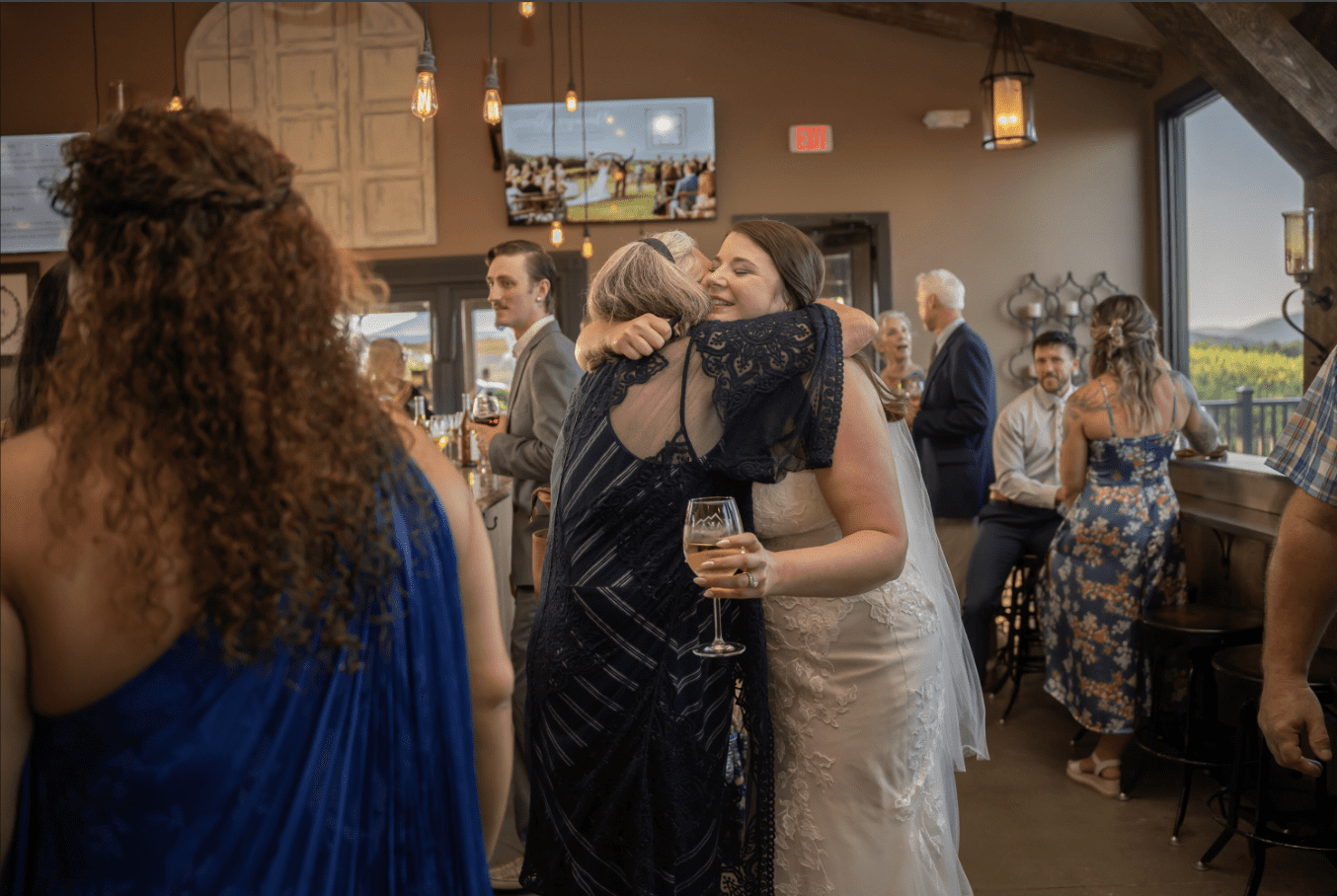 Bride hugging a woman, both smiling, inside a venue with guests.