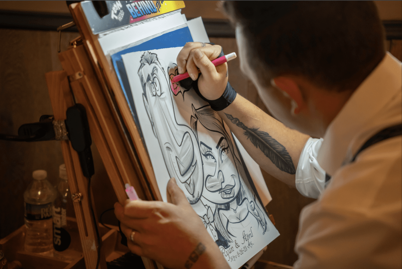 Artist drawing a caricature on an easel, using a pen. He is indoors, and a feather tattoo is visible.