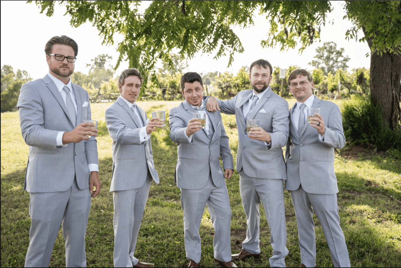 Five men in gray suits toasting with drinks outdoors.
