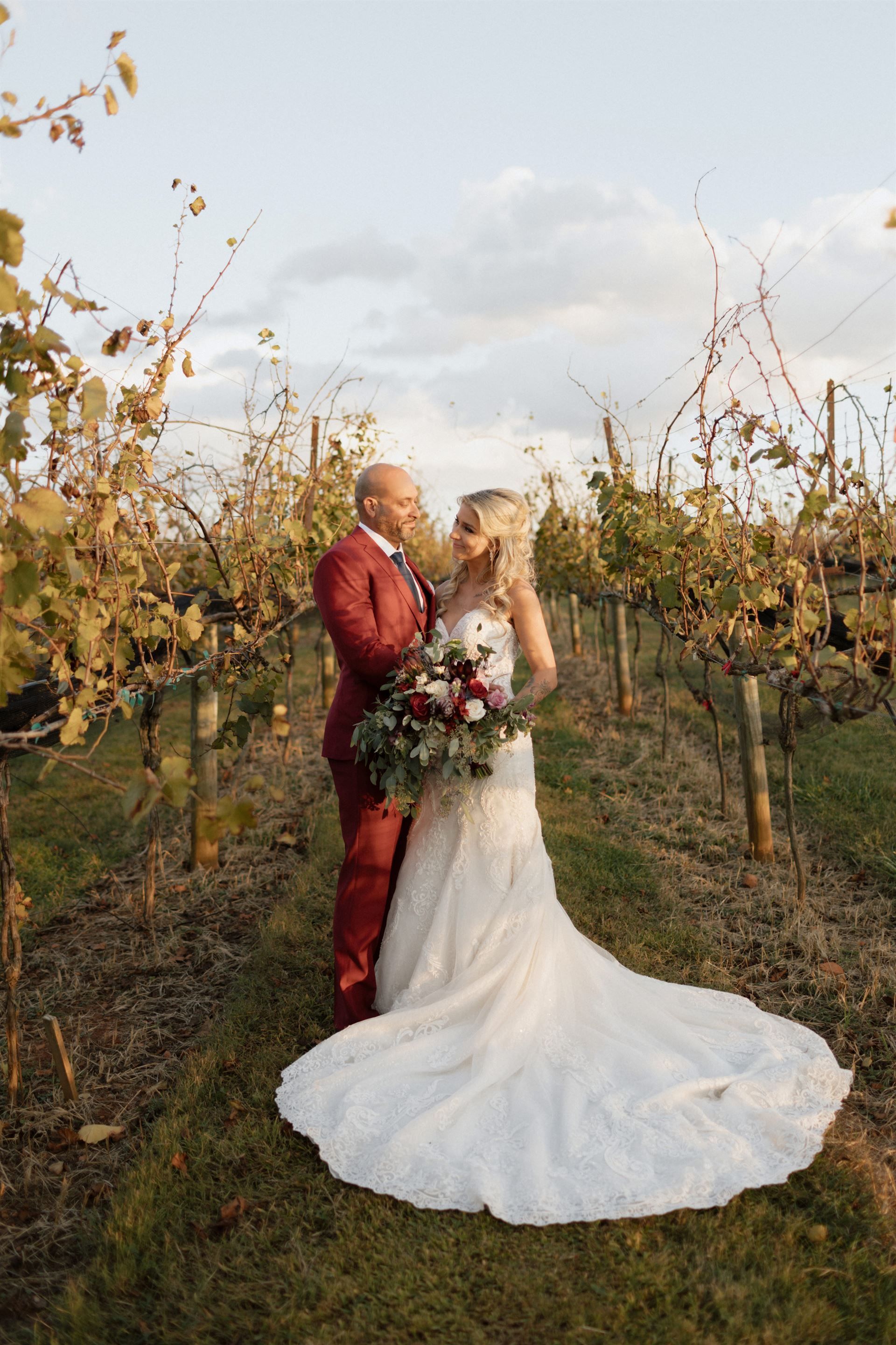 Bride and groom embrace in a vineyard. The groom wears a red suit, the bride a white gown, holding a bouquet.
