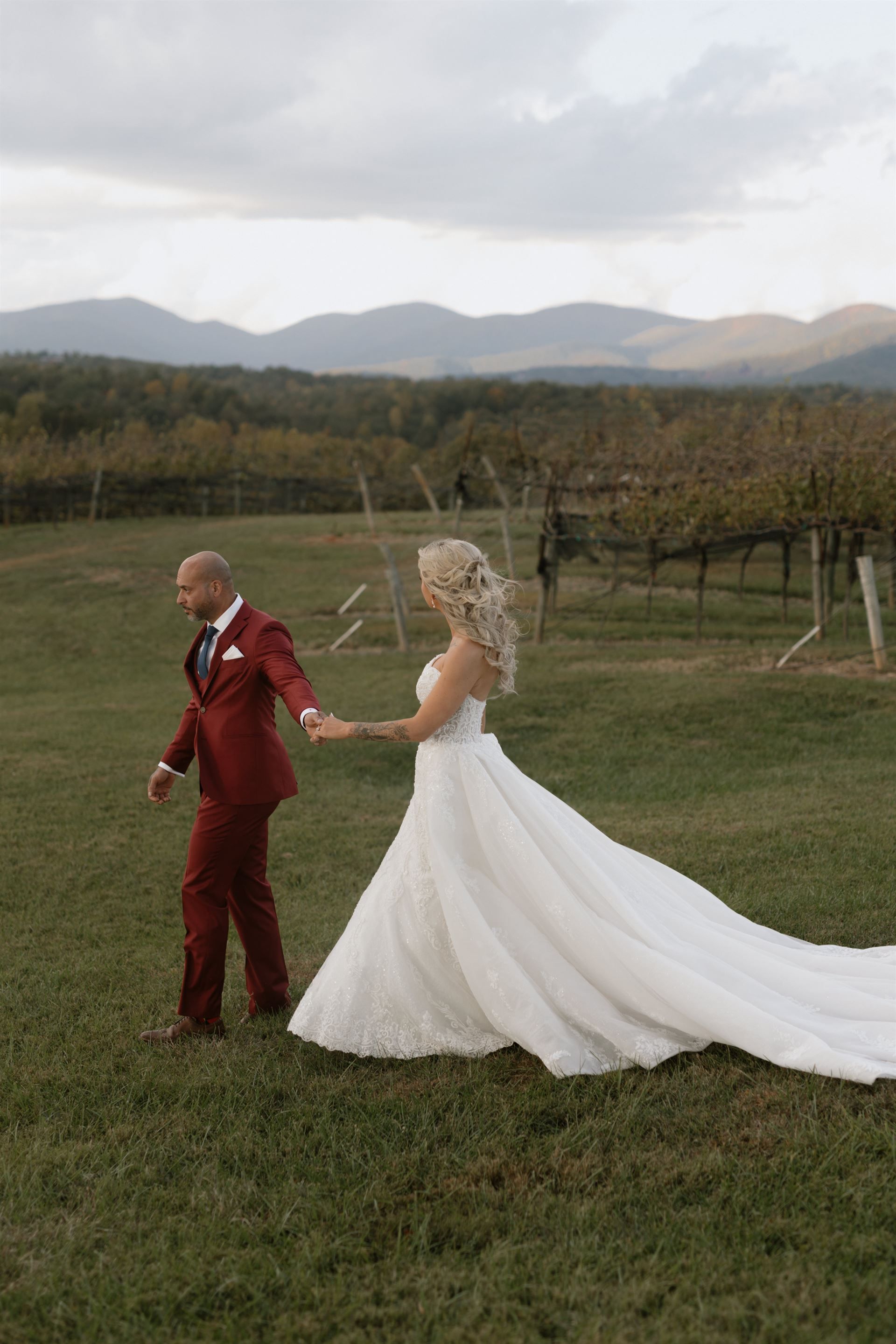 Couple in wedding attire walk hand-in-hand in a field, vineyard in background, mountains, red suit, white dress.