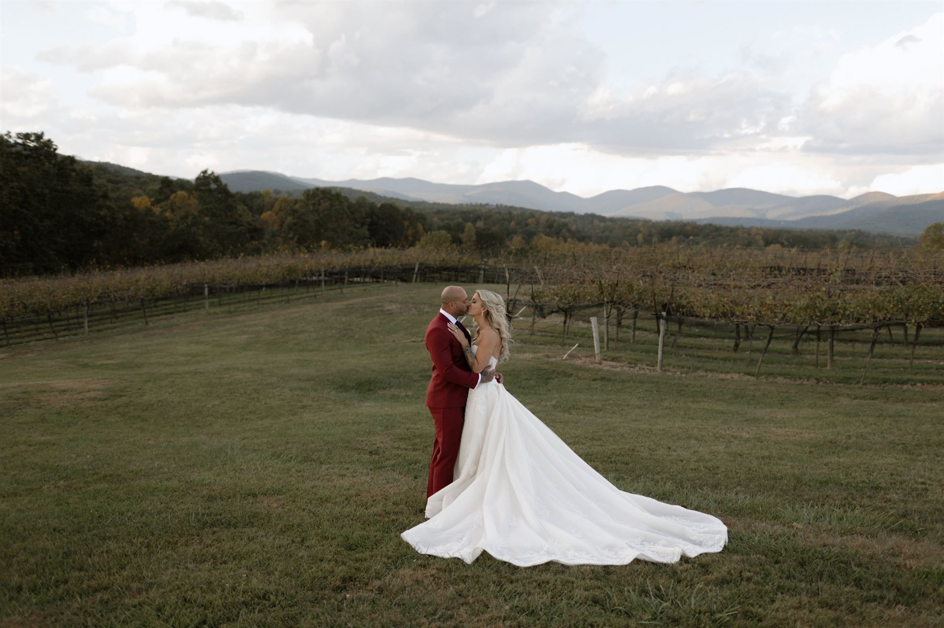 Couple embraces in vineyard, mountain backdrop. Groom in red suit, bride in white dress.