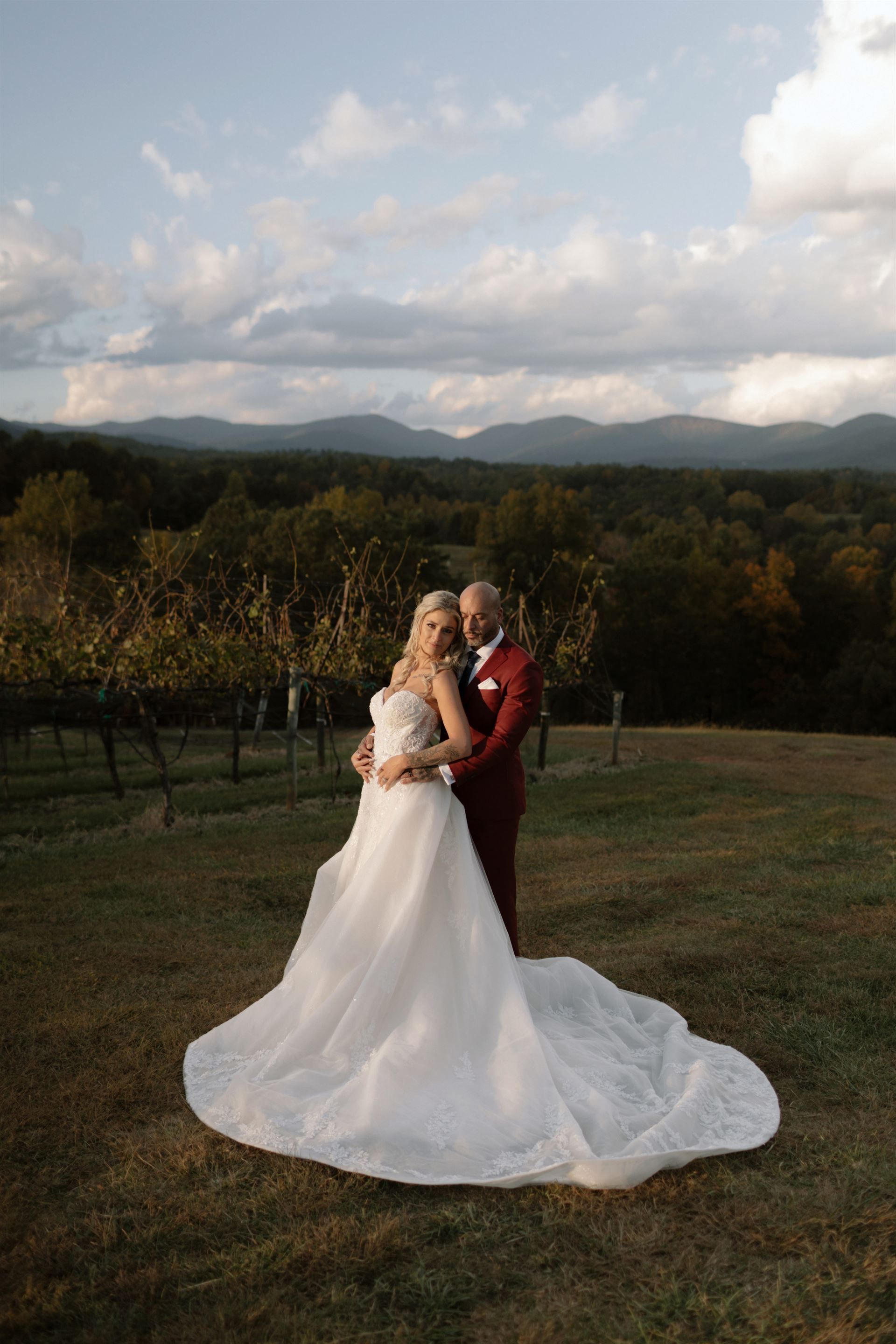 Bride and groom embrace on a hill, overlooking a vineyard and mountains at sunset.