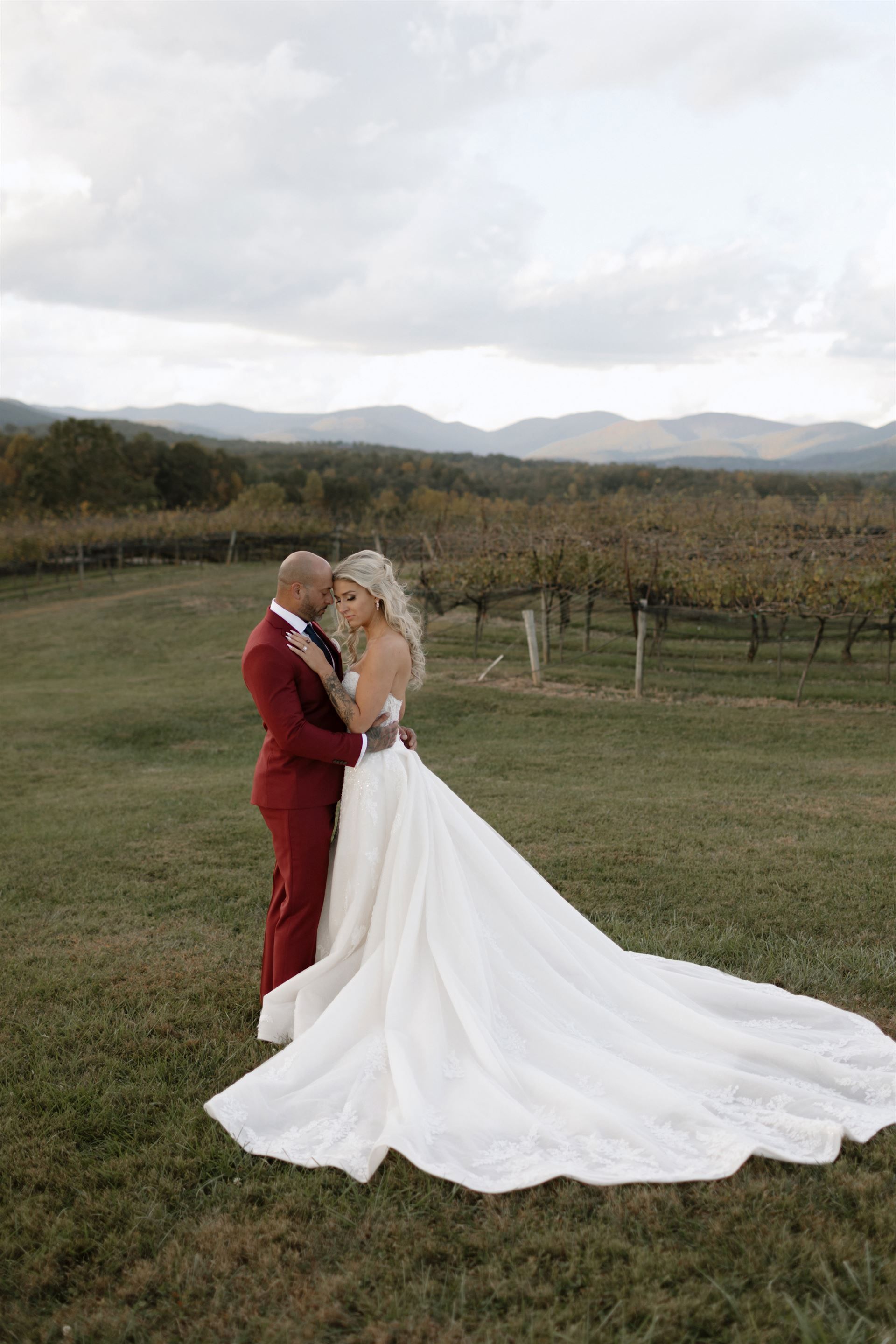 Couple embraces at a vineyard, groom in red suit, bride in white gown, mountains in background.
