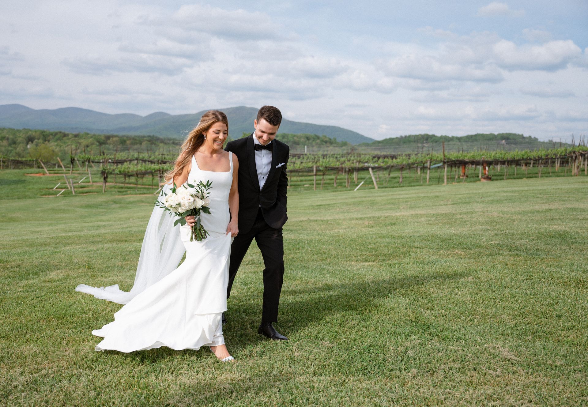 Bride and groom walk hand-in-hand in a grassy field, mountains in the background, vineyard visible.