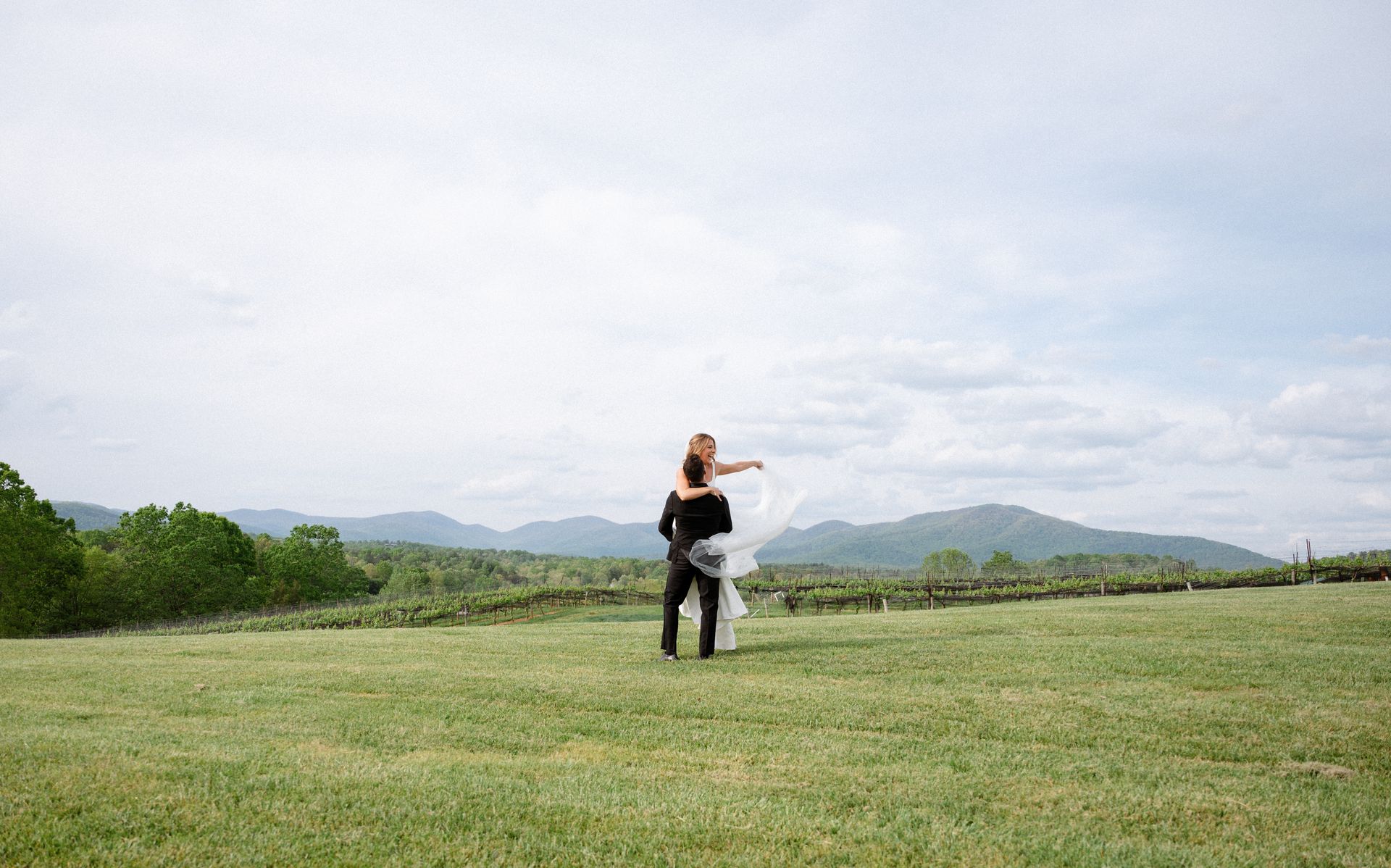 Couple dancing on a grassy field with mountains in the background under a cloudy sky.