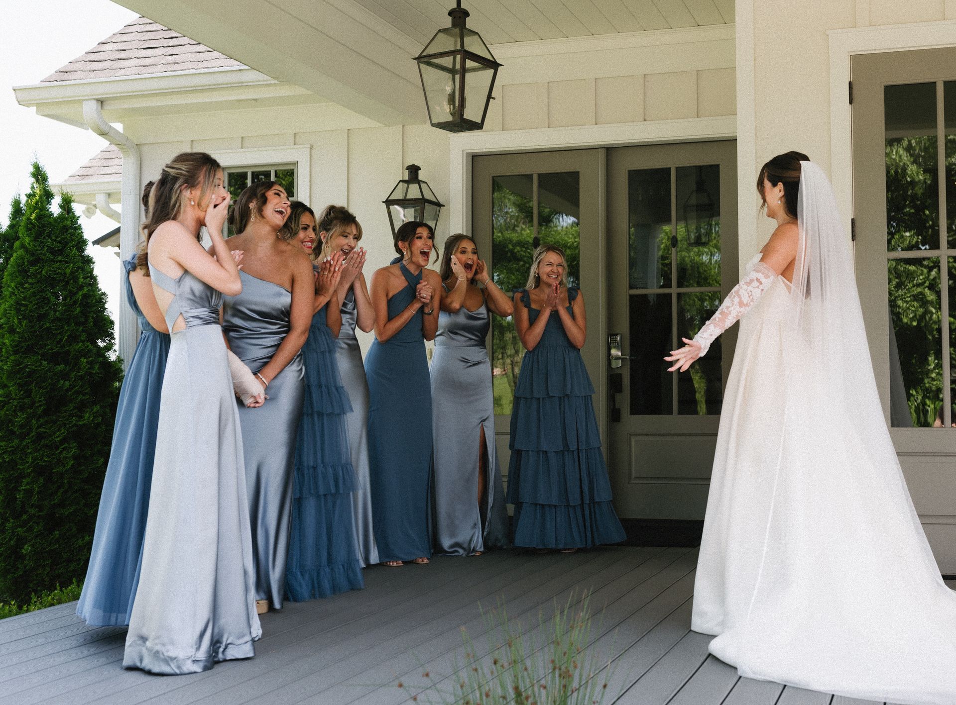 Bride reveals herself to bridesmaids on a porch; they react with surprise and joy.