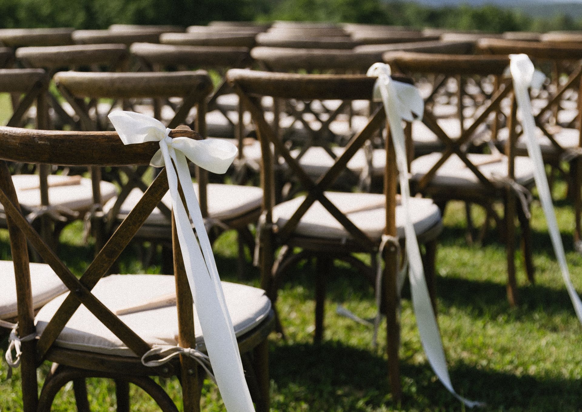Wooden chairs with cream cushions and white ribbon decorations arranged outdoors on a grassy field.