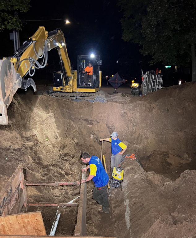 Two men are working in a dirt pit at night with a bulldozer in the background