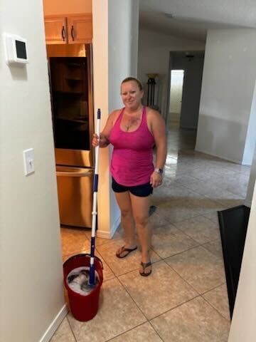 Woman in a pink tank top stands in a tiled hallway holding a mop beside a red bucket.