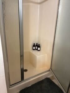 Shower stall with two small bottles on a built-in shelf and a black bath mat below.