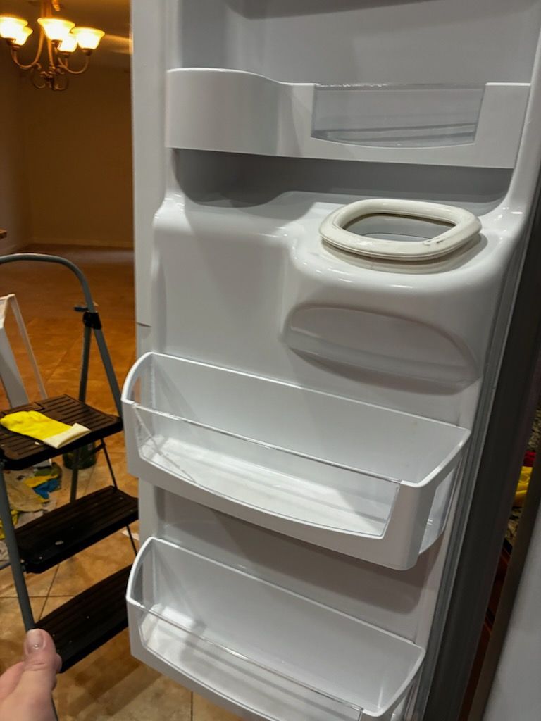 Interior of an empty refrigerator door with white shelves. A step ladder is in the background.