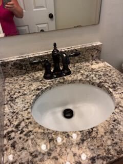Bathroom sink with speckled granite countertop, black faucet, and a mirror reflecting part of a person in pink.