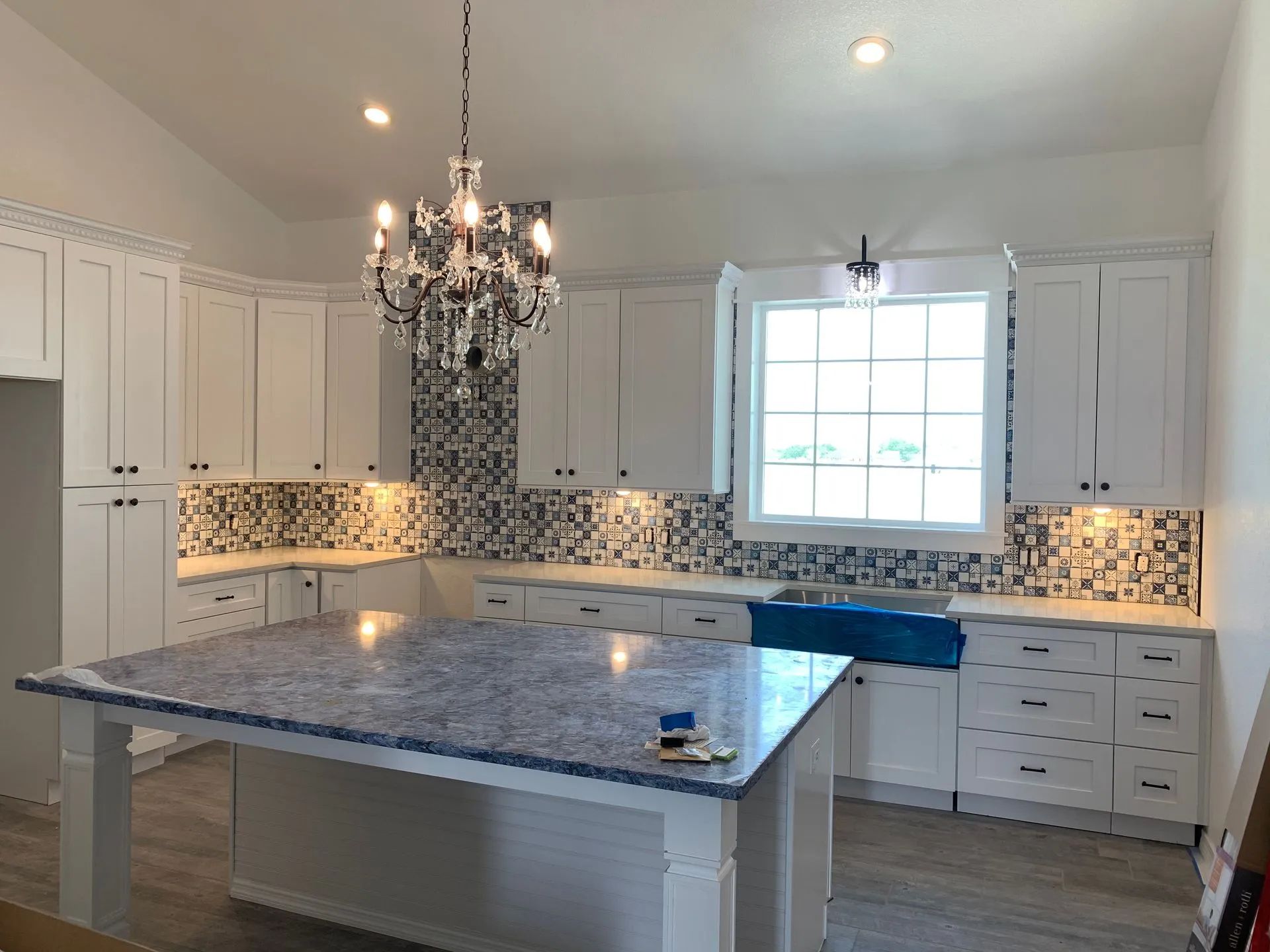 Bright white kitchen with blue island countertop, chandelier, mosaic backsplash, and large window