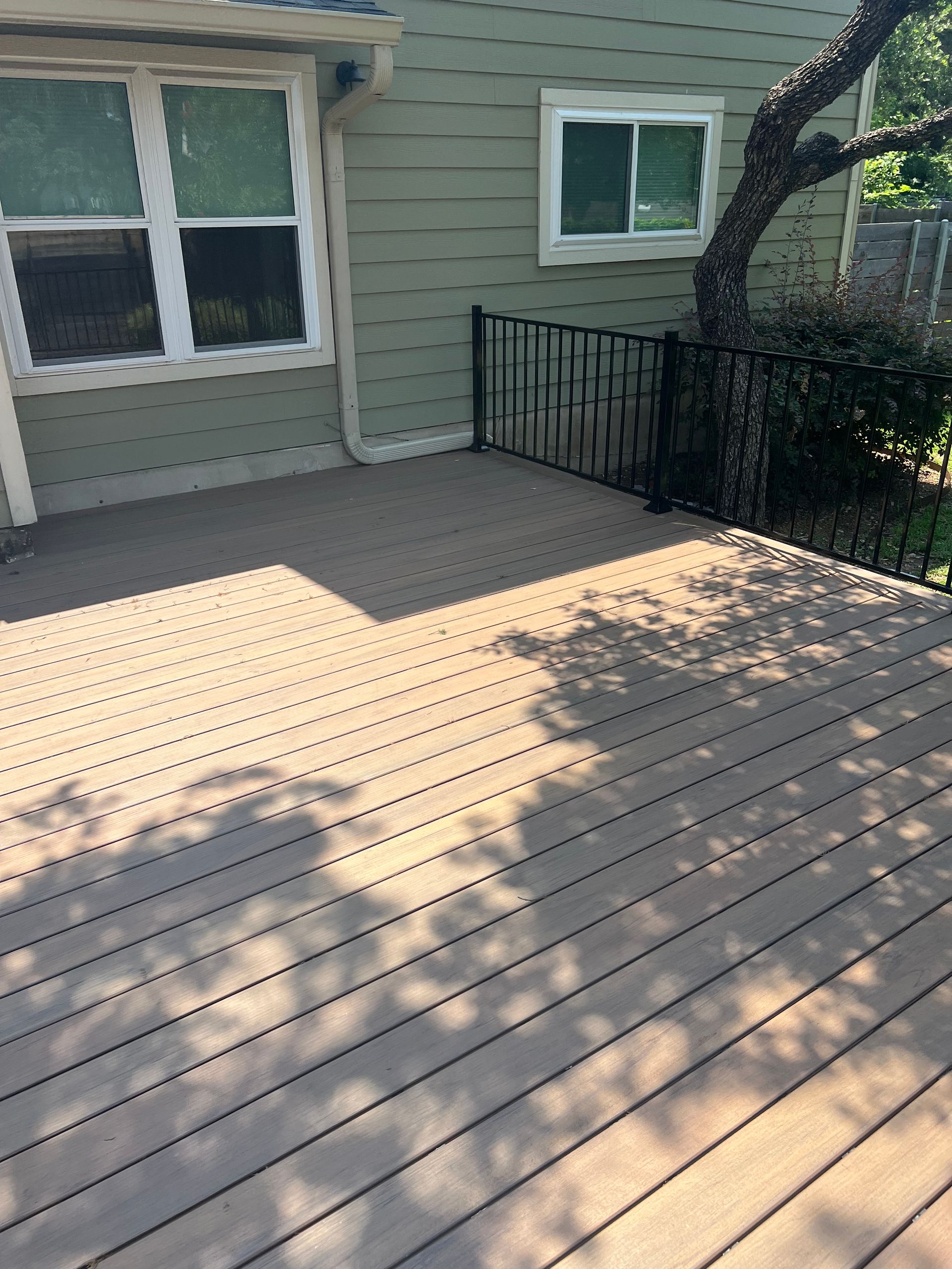 Wooden deck patio beside a house with green siding, railings, and tree shadows on the boards.