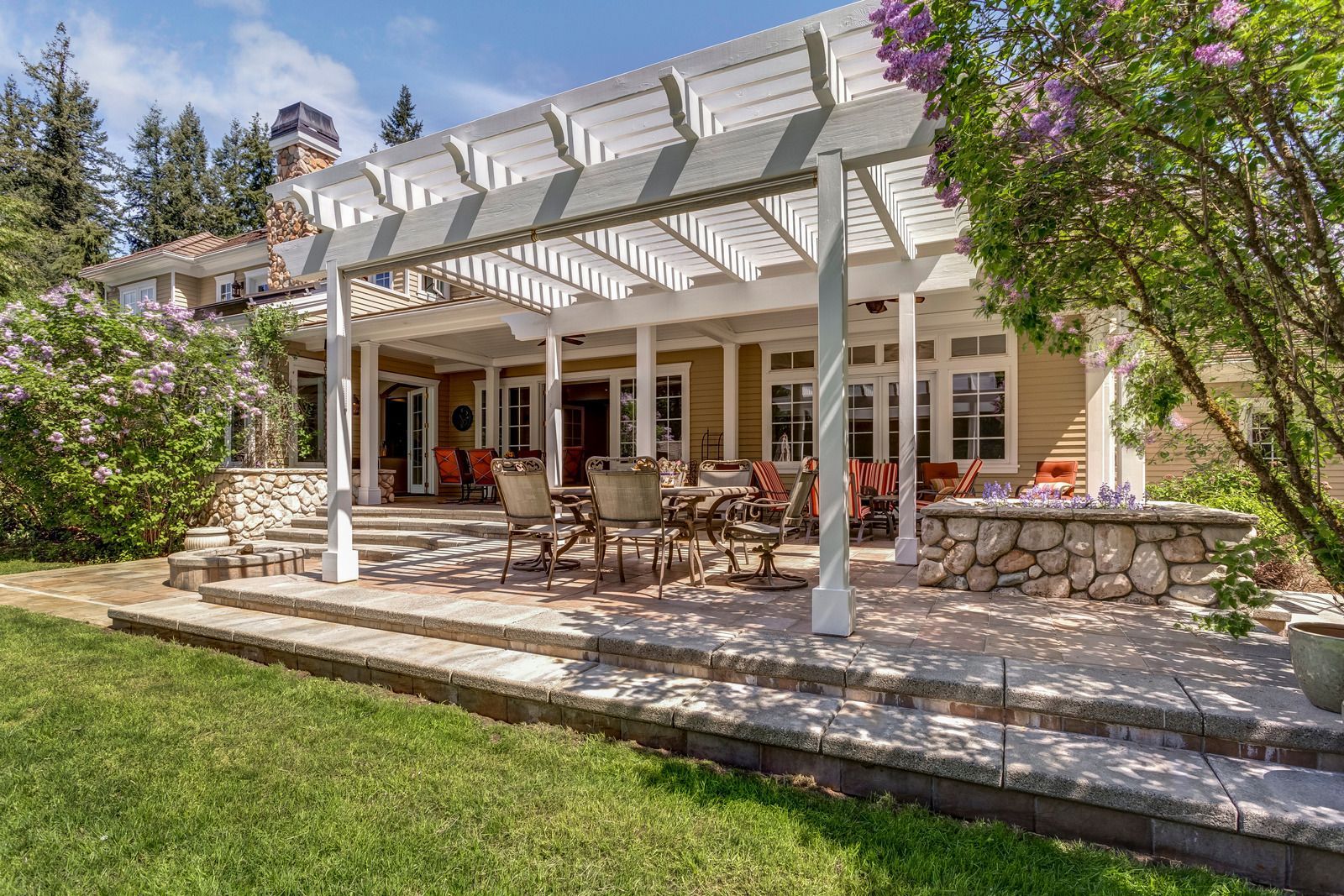 Backyard patio with pergola, stone steps, table, chairs, and tan house under a sunny sky.