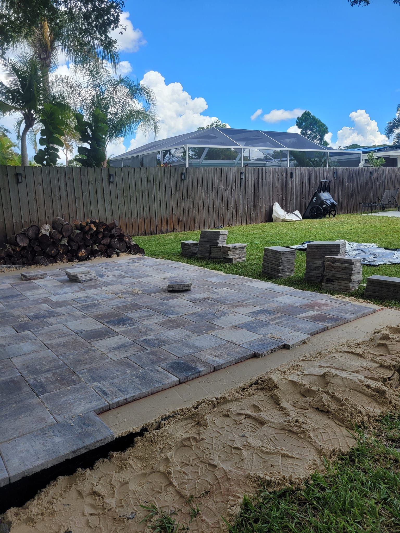 Patio pavers being installed in a backyard with a wooden fence and blue sky.