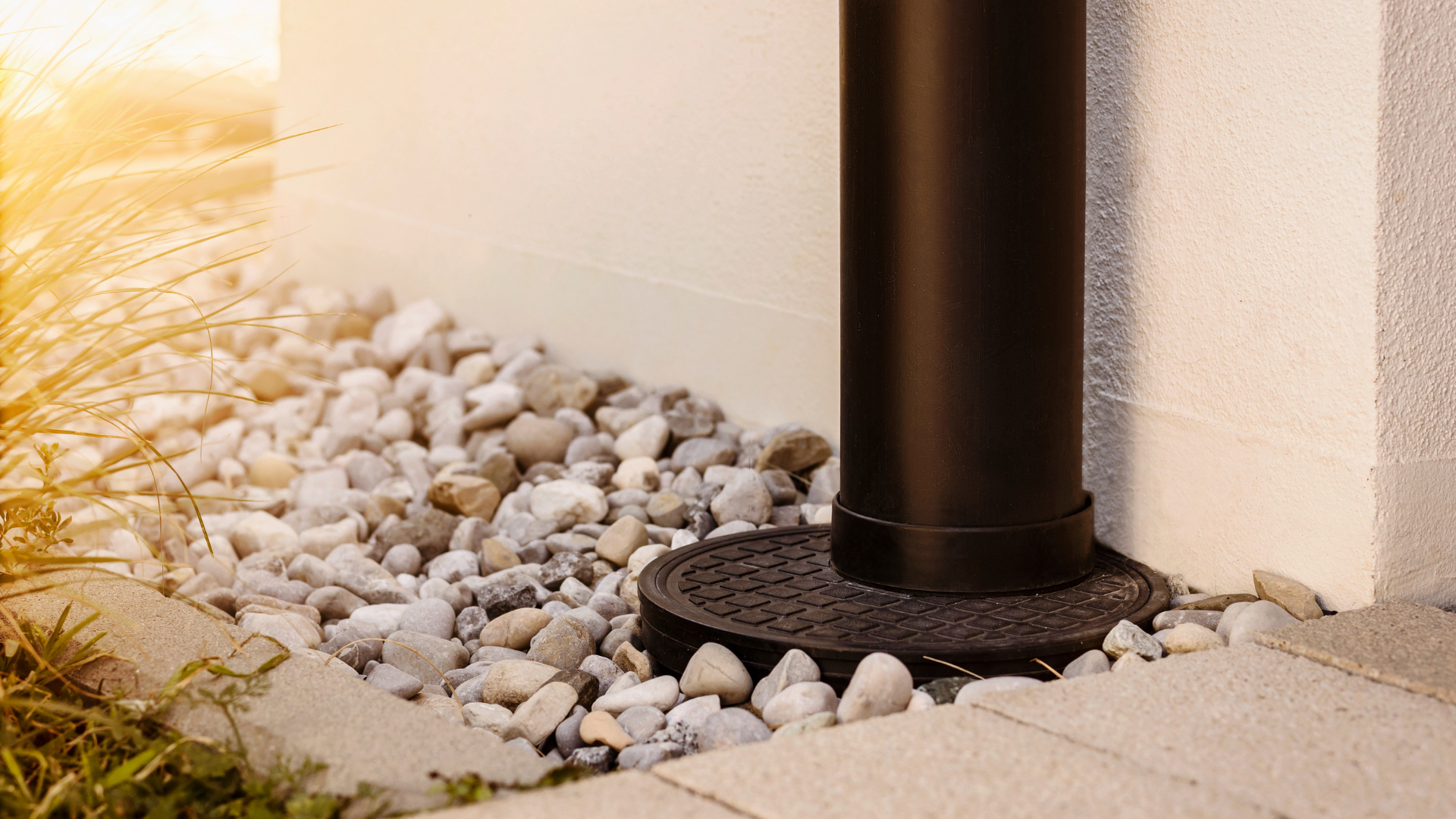 Black pipe against a white wall with a decorative gravel base. Sunlight.