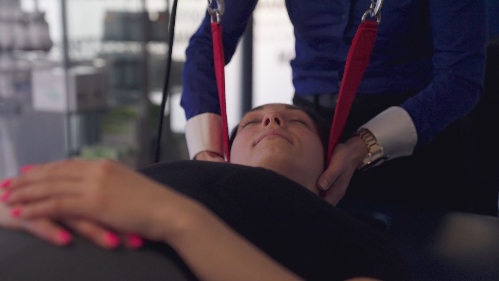 Woman receiving neck traction from a medical professional. Red straps, closed eyes, indoor setting.