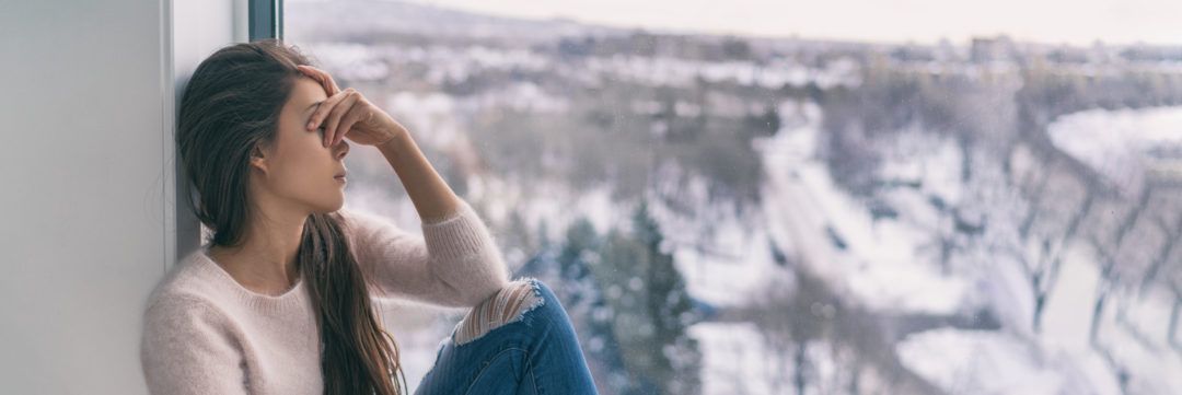 Woman sitting by a window, looking pensive. Snowy landscape visible outside.