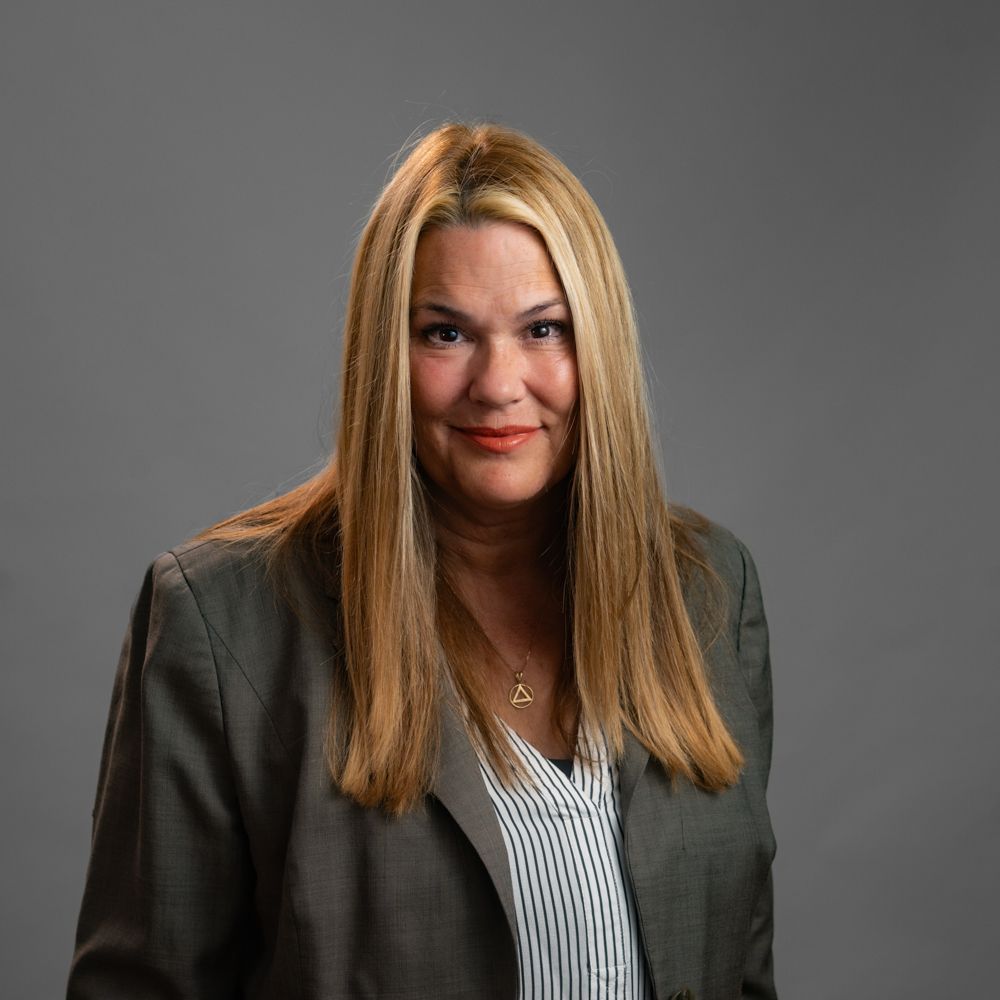 Woman with long blonde hair wearing a gray suit jacket and striped shirt, smiling, against a gray backdrop.