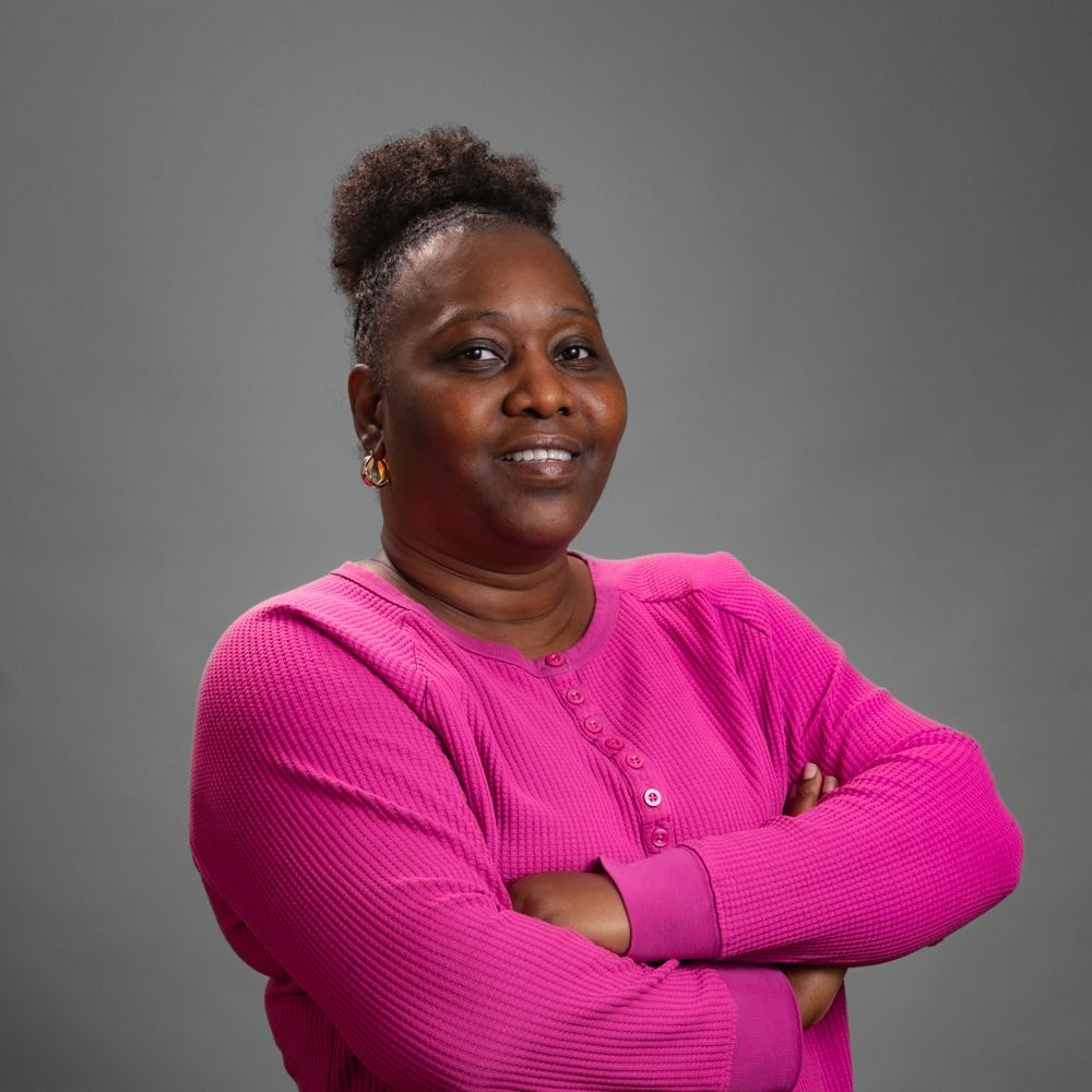 Woman with arms crossed, wearing pink top, against gray background.
