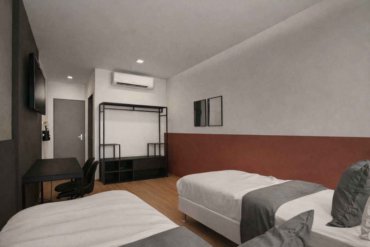 Um quarto de hotel minimalista com duas camas de solteiro brancas, uma escrivaninha preta e um guarda-roupa aberto de metal encostado em uma parede de dois tons.