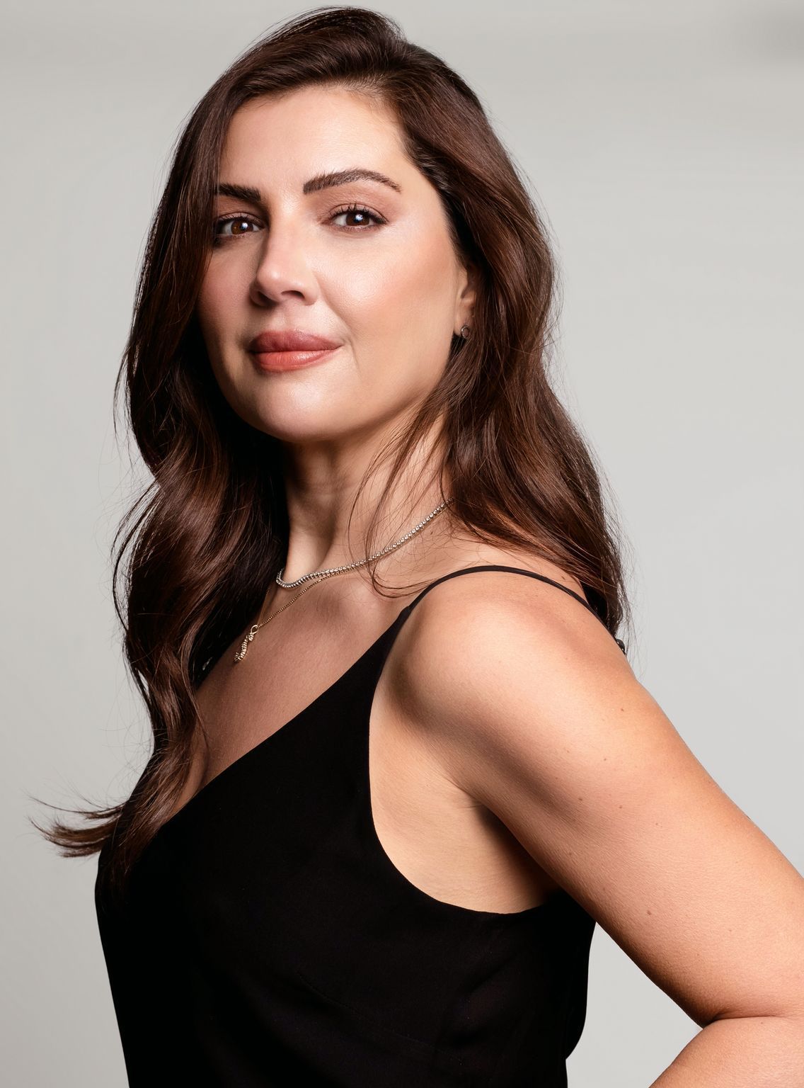 Woman with long, wavy brown hair wearing a black spaghetti strap top and gold necklace, smiling, neutral background.