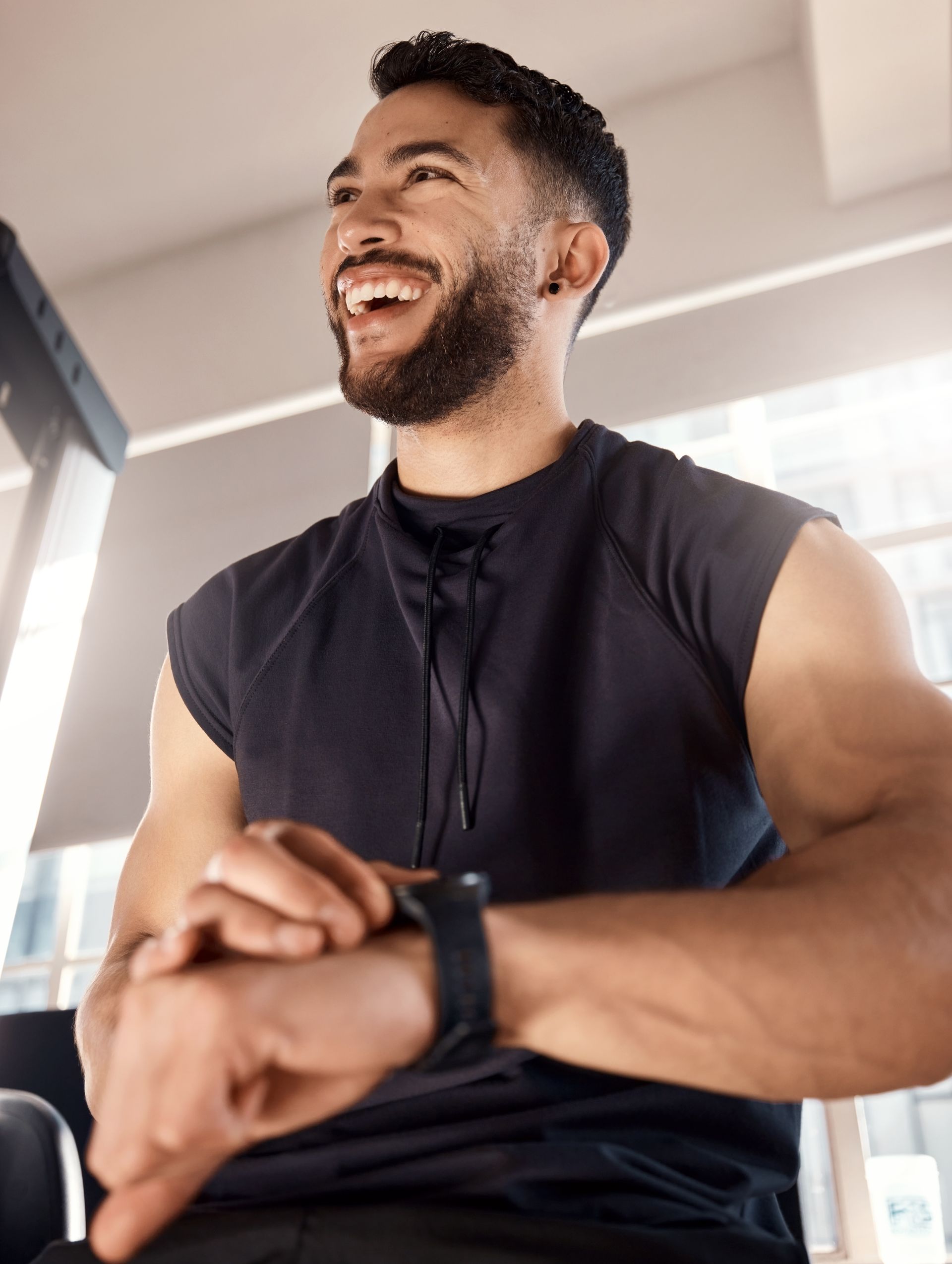 Man smiles, checks watch in a gym setting. Wearing black sleeveless shirt.