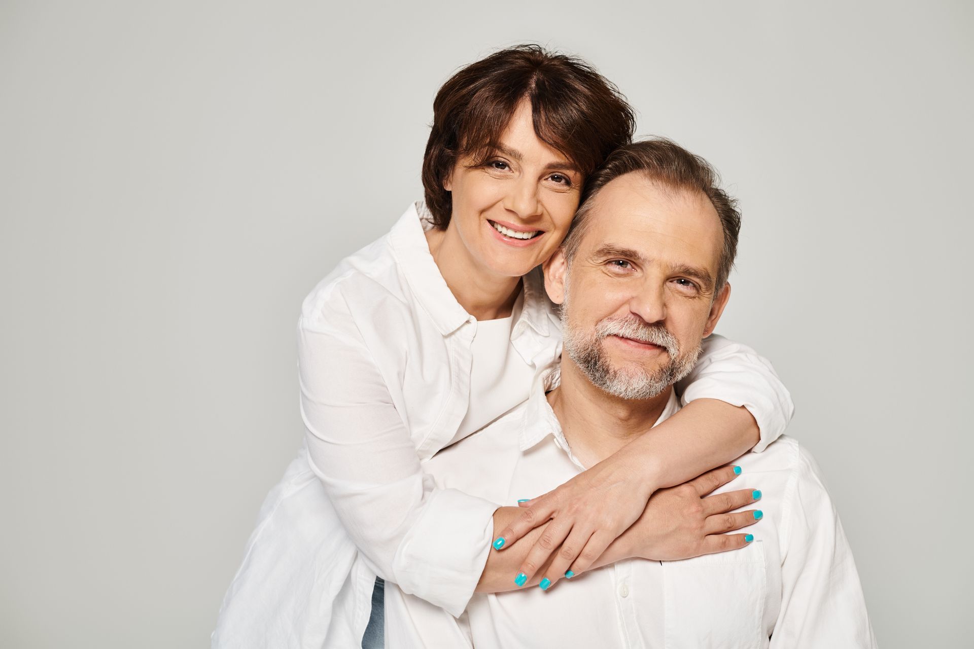 Woman hugging a man from behind, both smiling, wearing white shirts, against a light gray background.