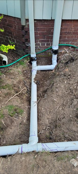 PVC pipes and hoses installed along a brick wall over bare soil in a yard