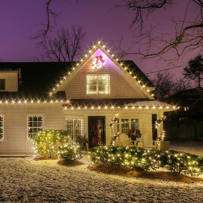 House decorated with warm white holiday lights at dusk, with a glowing purple sky and snow-covered yard