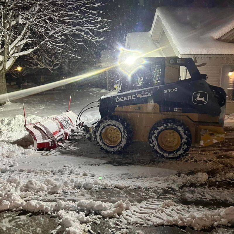 Skid steer with red snow blower clearing a snowy driveway at night beside a house