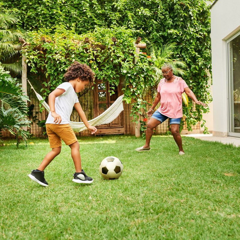 Two people playing soccer on a grassy backyard lawn near a hammock and ivy-covered wall